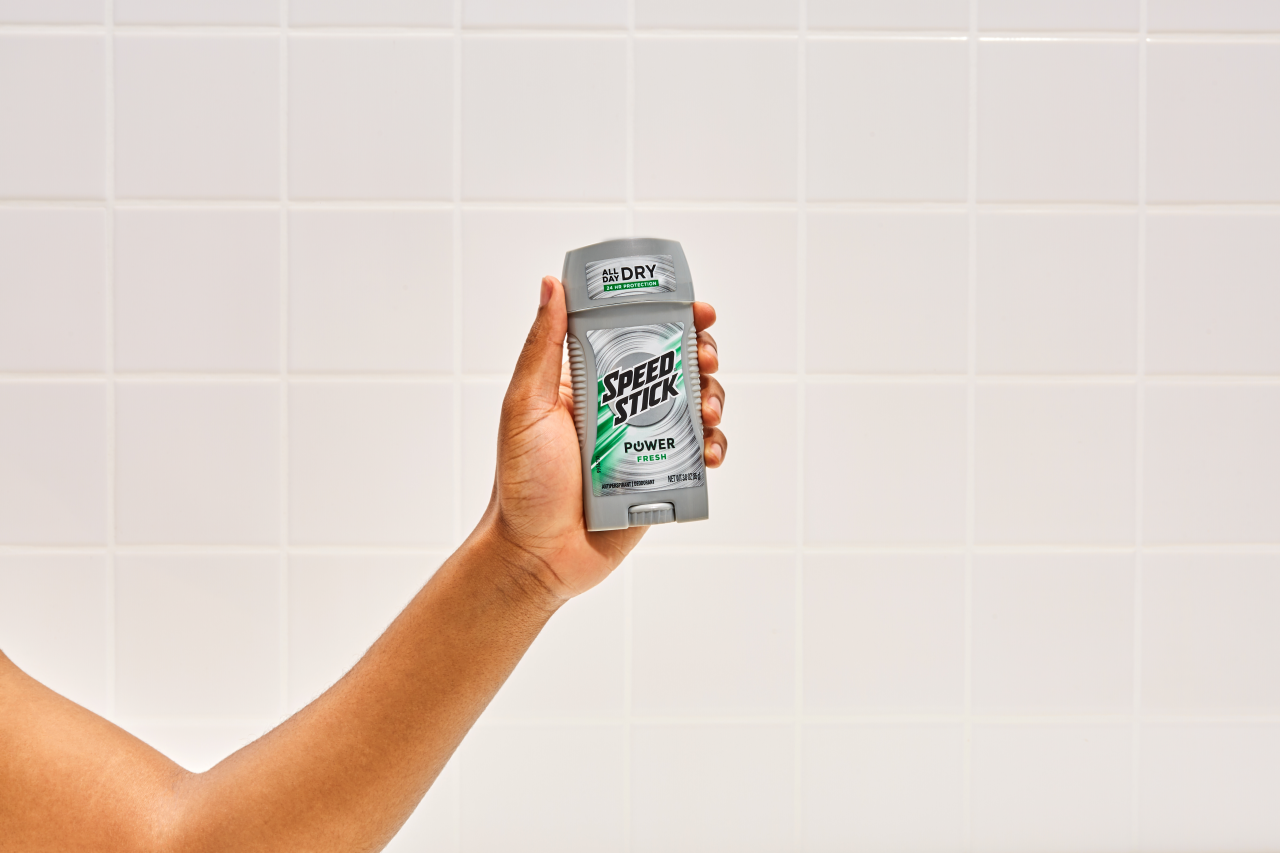 Hand holding Speed Stick antiperspirant deodorant stick against tiled bathroom wall.