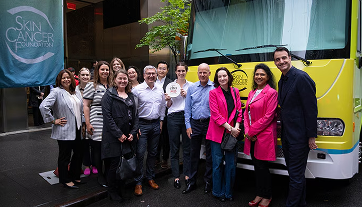 Group of people smiling at the camera while standing in front of a yellow vehicle as part of an event for "Skin Cancer Foundation".