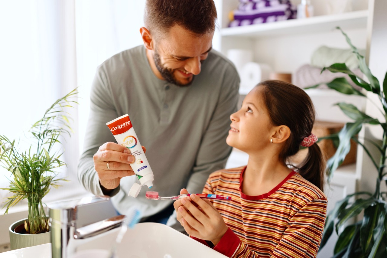 A man applying toothpaste to a girl's toothbrush at a sink, with plants in the background.